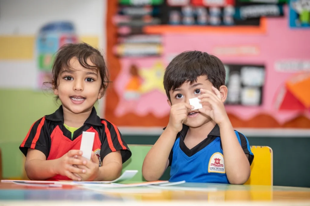 Two preschool children in colorful uniforms participating in hands-on learning activities in a vibrant classroom, smiling and exploring materials.