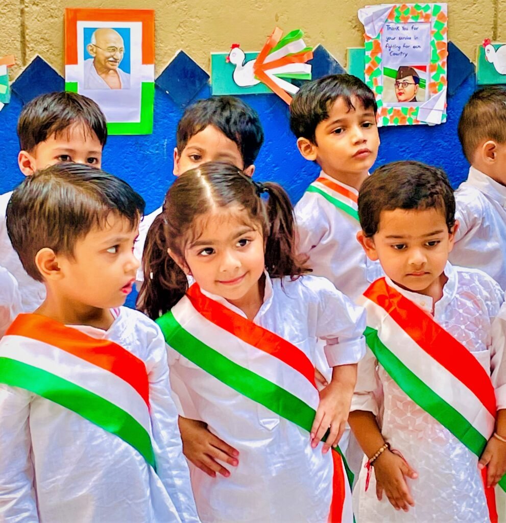 Group of young children dressed in white traditional attire with tricolor sashes, celebrating Independence Day with patriotic decorations and portraits of national leaders in the background.