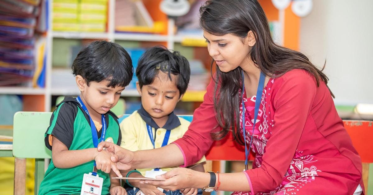 A young teacher in a red dress guiding two preschool children during a learning activity in a colorful classroom, fostering interactive education.