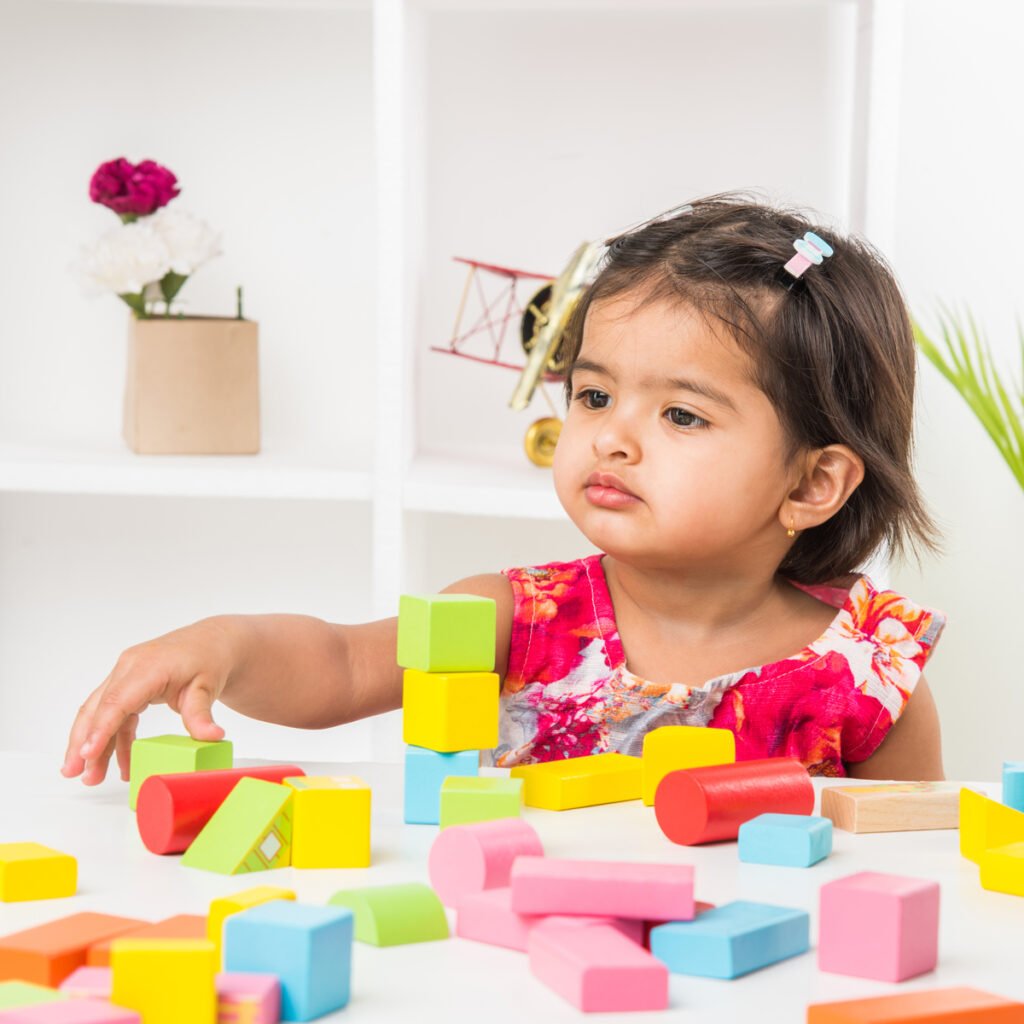 A young child in a vibrant floral dress stacking colorful building blocks on a white table, with decorative shelves in the background.