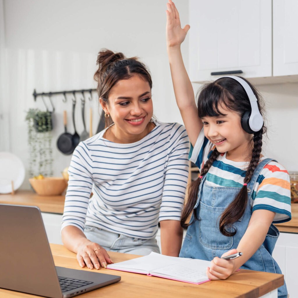 A young girl wearing headphones raising her hand enthusiastically while studying with a laptop and notebook, supported by her smiling mother in a cozy home