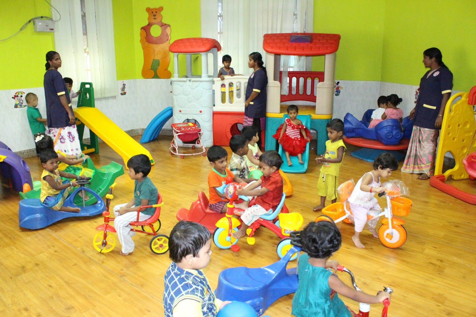 A vibrant indoor play area with children riding tricycles, playing on slides, and enjoying various activities, supervised by caregivers in a colorful setting.