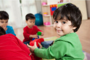 A cheerful toddler wearing a green shirt playing with colorful building blocks in a preschool classroom, with other children playing in the background.