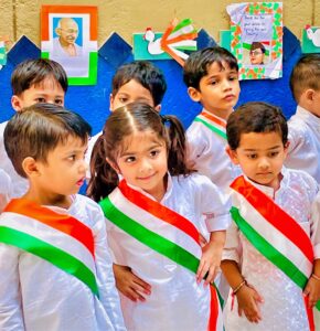 A group of children dressed in white outfits with tricolor sashes, celebrating Independence Day with patriotic decorations featuring Mahatma Gandhi and the Indian flag.